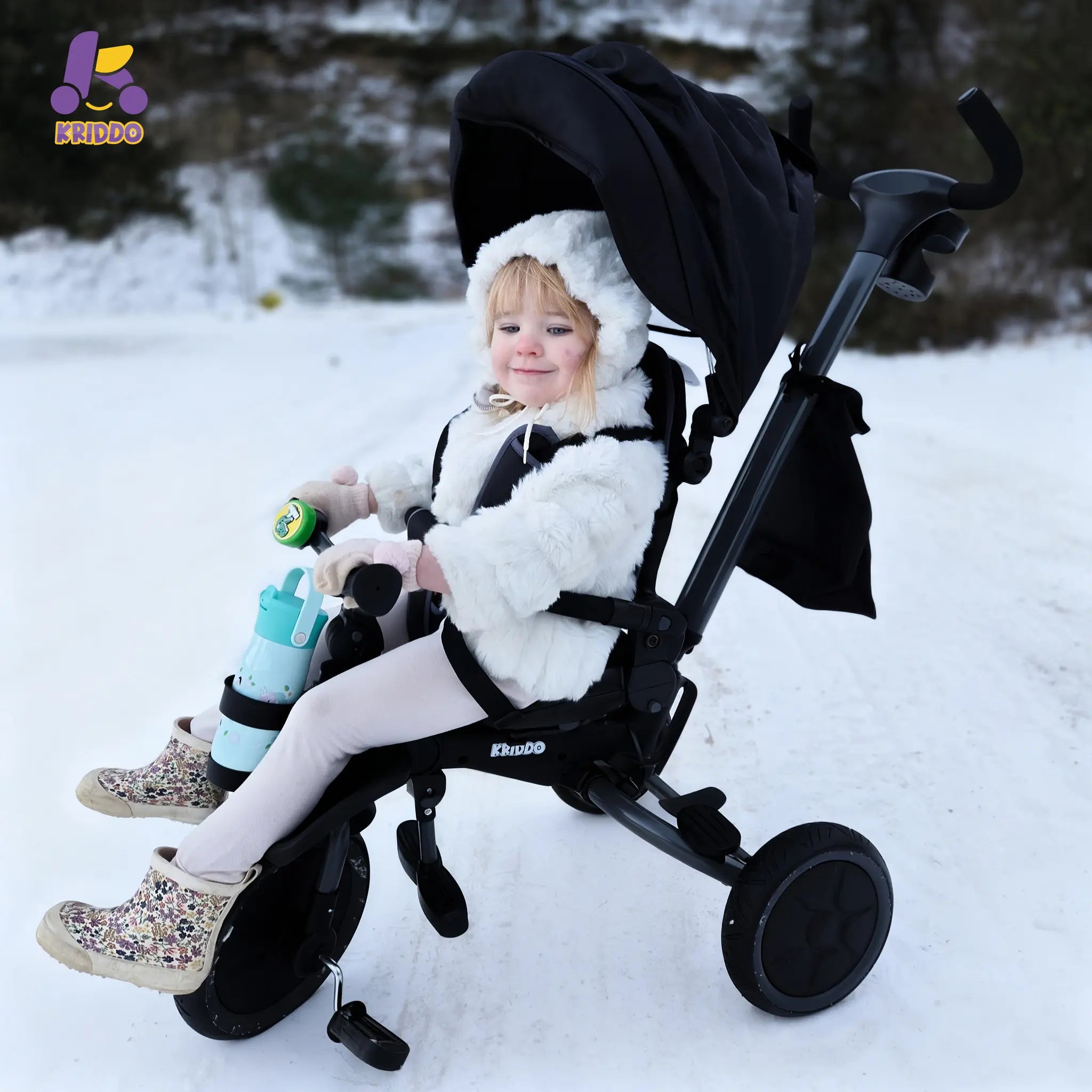Child in KRIDDO 8 in 1 tricycle stroller with a snowflake design in a snowy area.