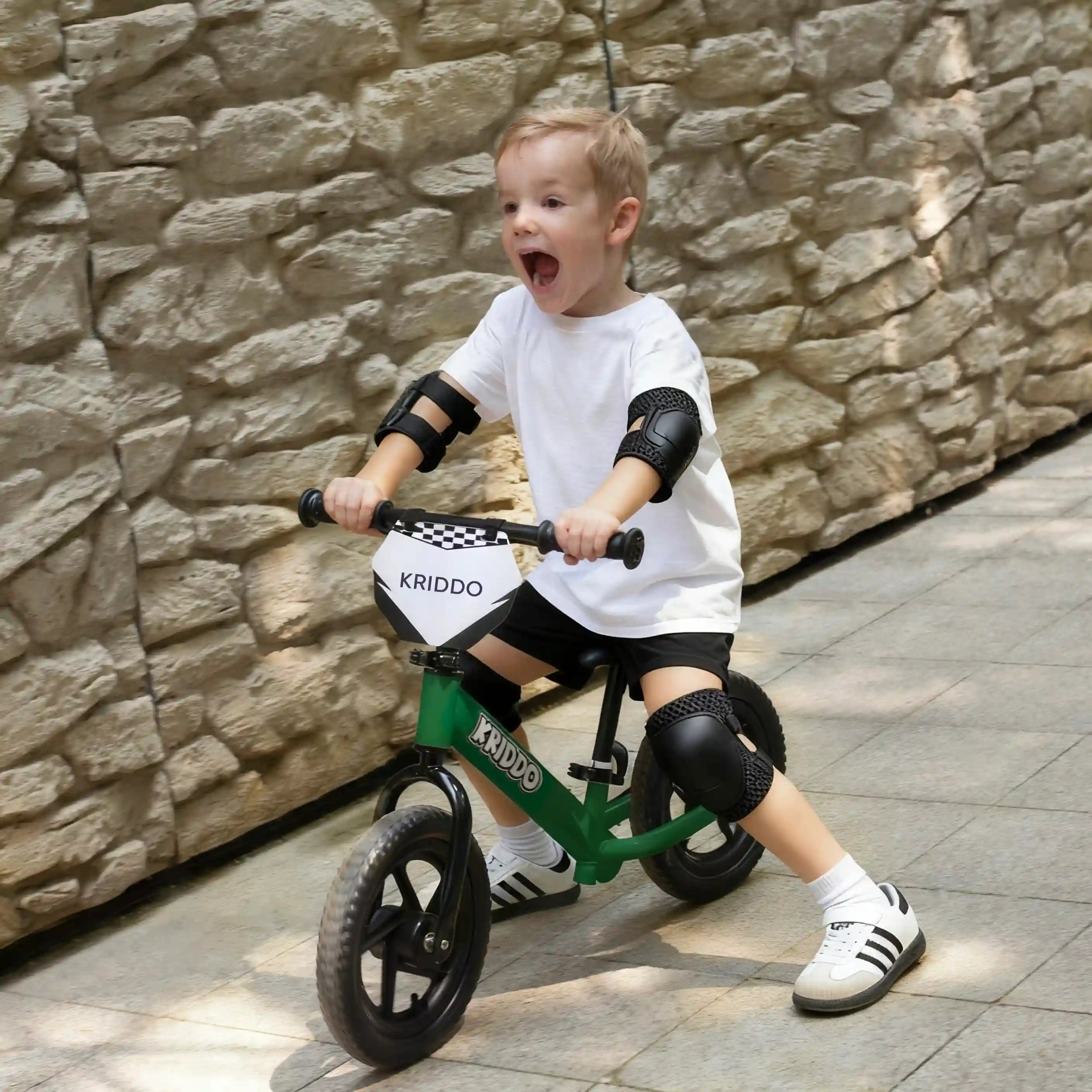 Child riding KRIDDO green 12 inch balance bike with elbow and knee pads.