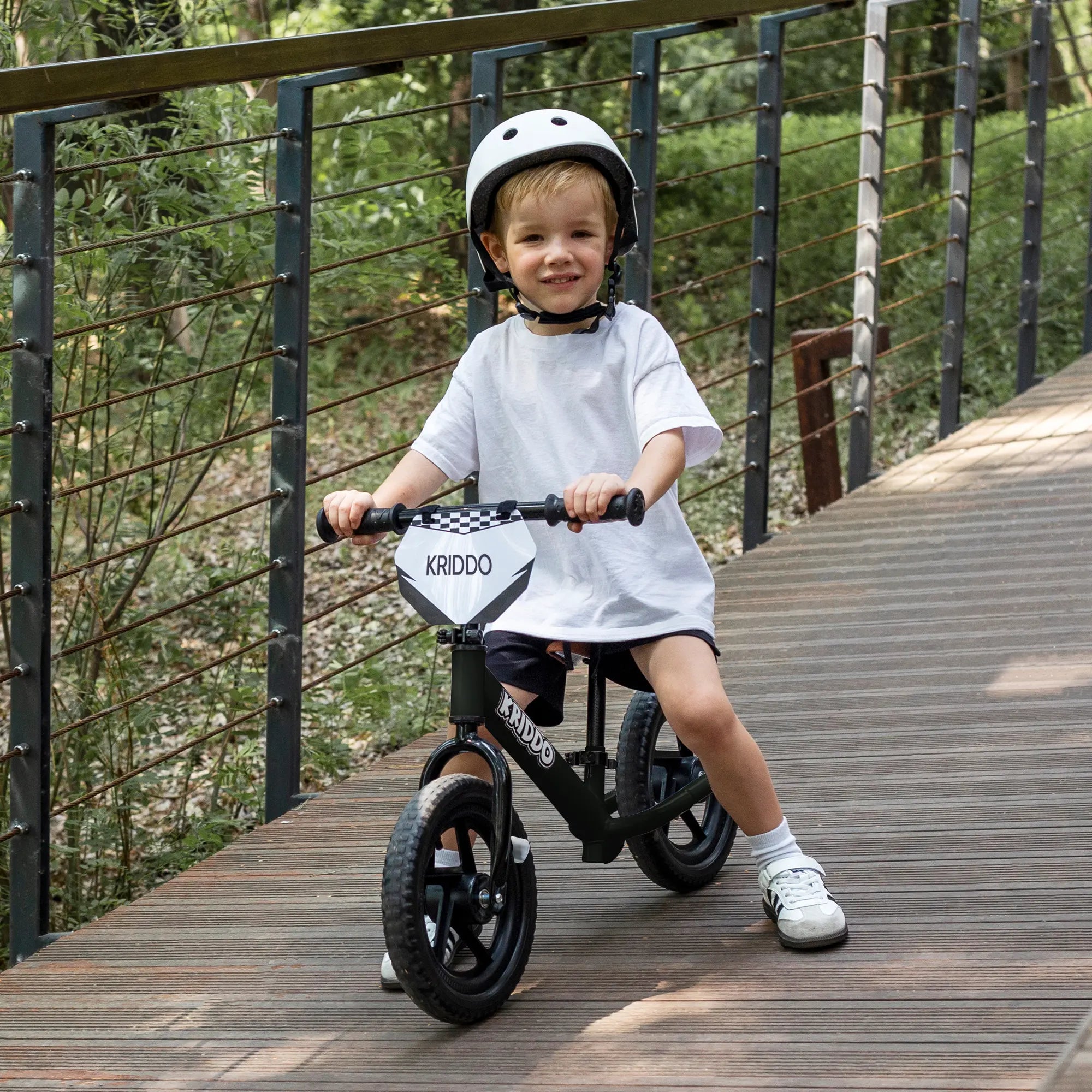 Toddler boy riding KRIDDO 12 inch balance bike on a wooden path with a helmet