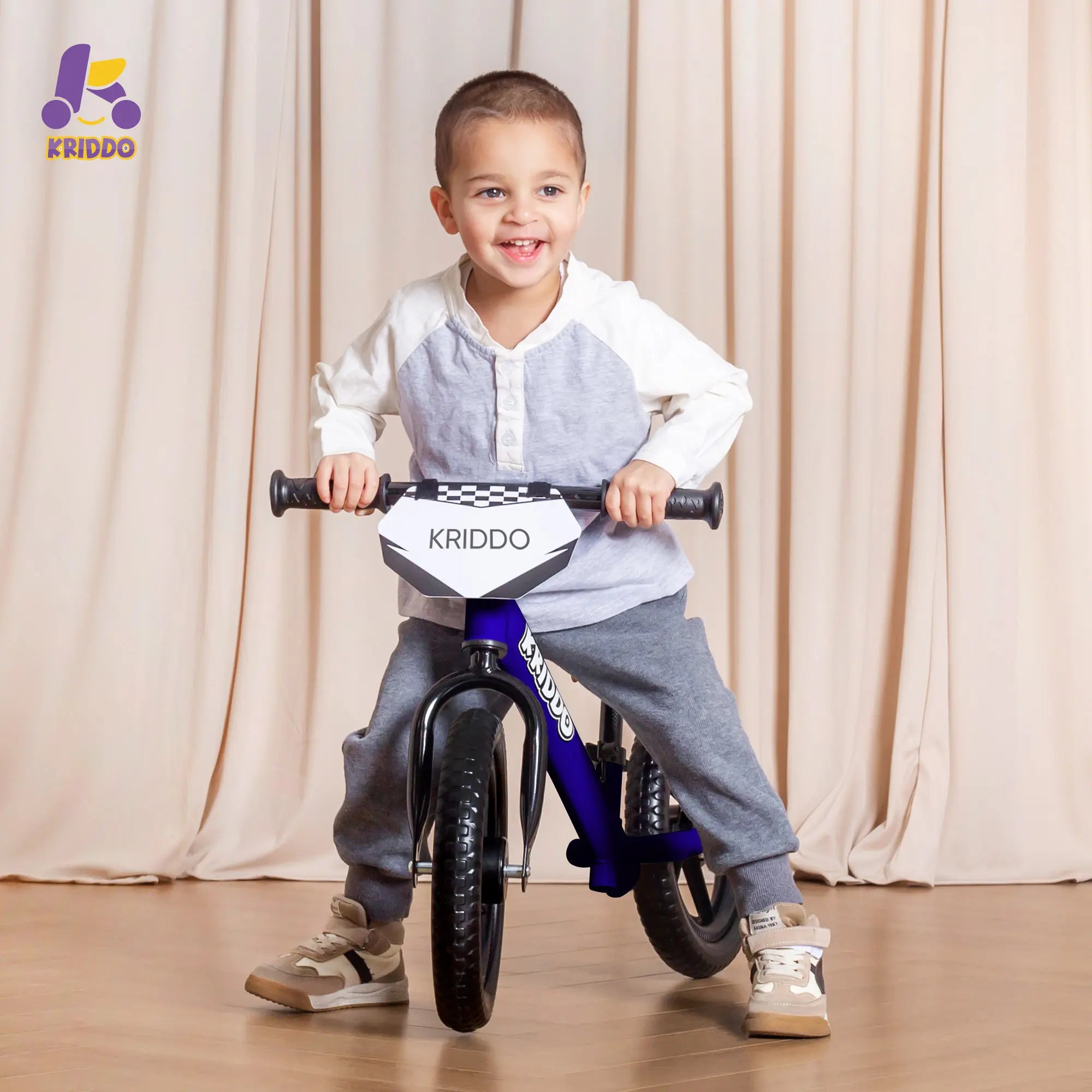 Toddler boy riding KRIDDO 12 Inch blue balance bike against a beige curtain