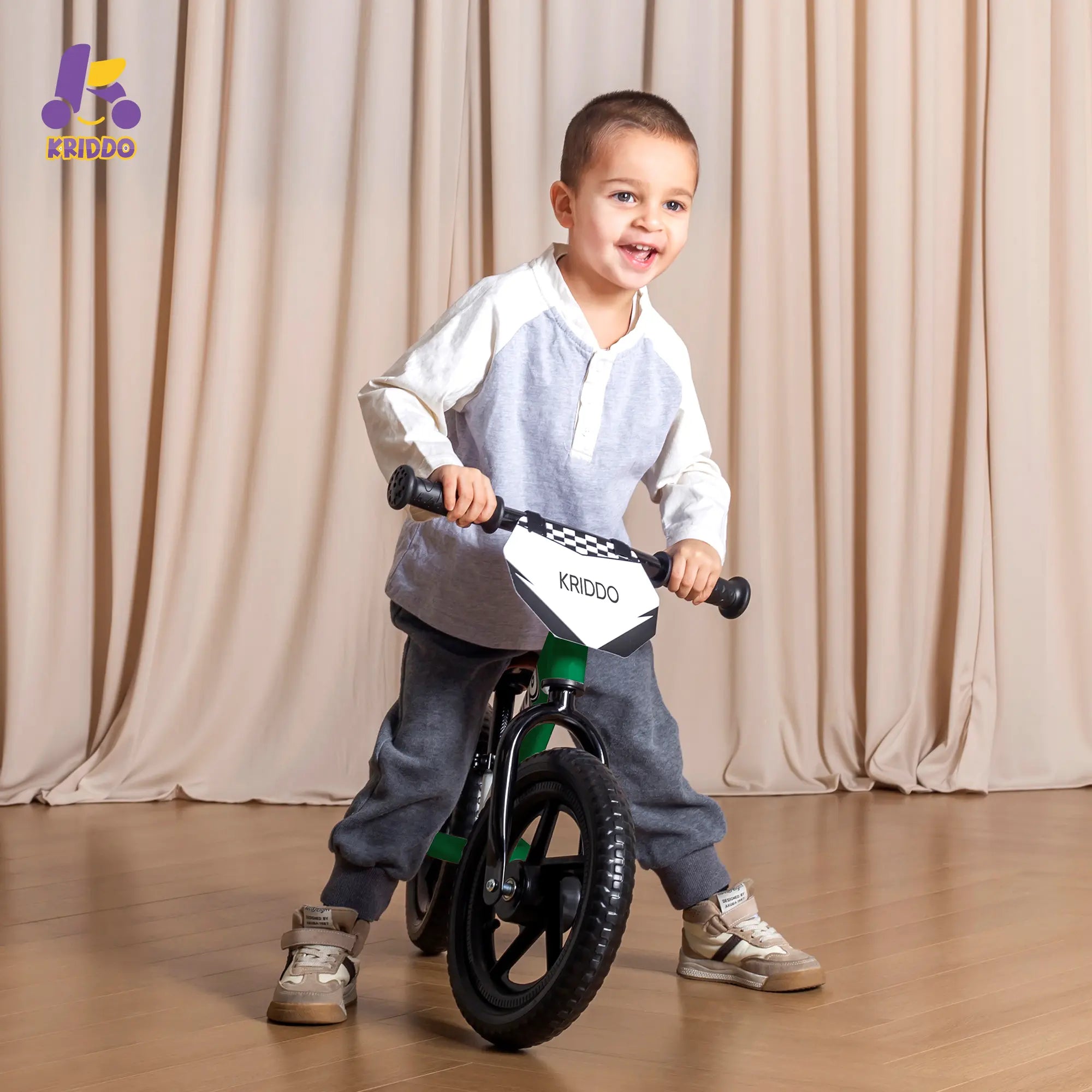 Toddler boy riding KRIDDO 12 Inch green balance bike against a beige curtain