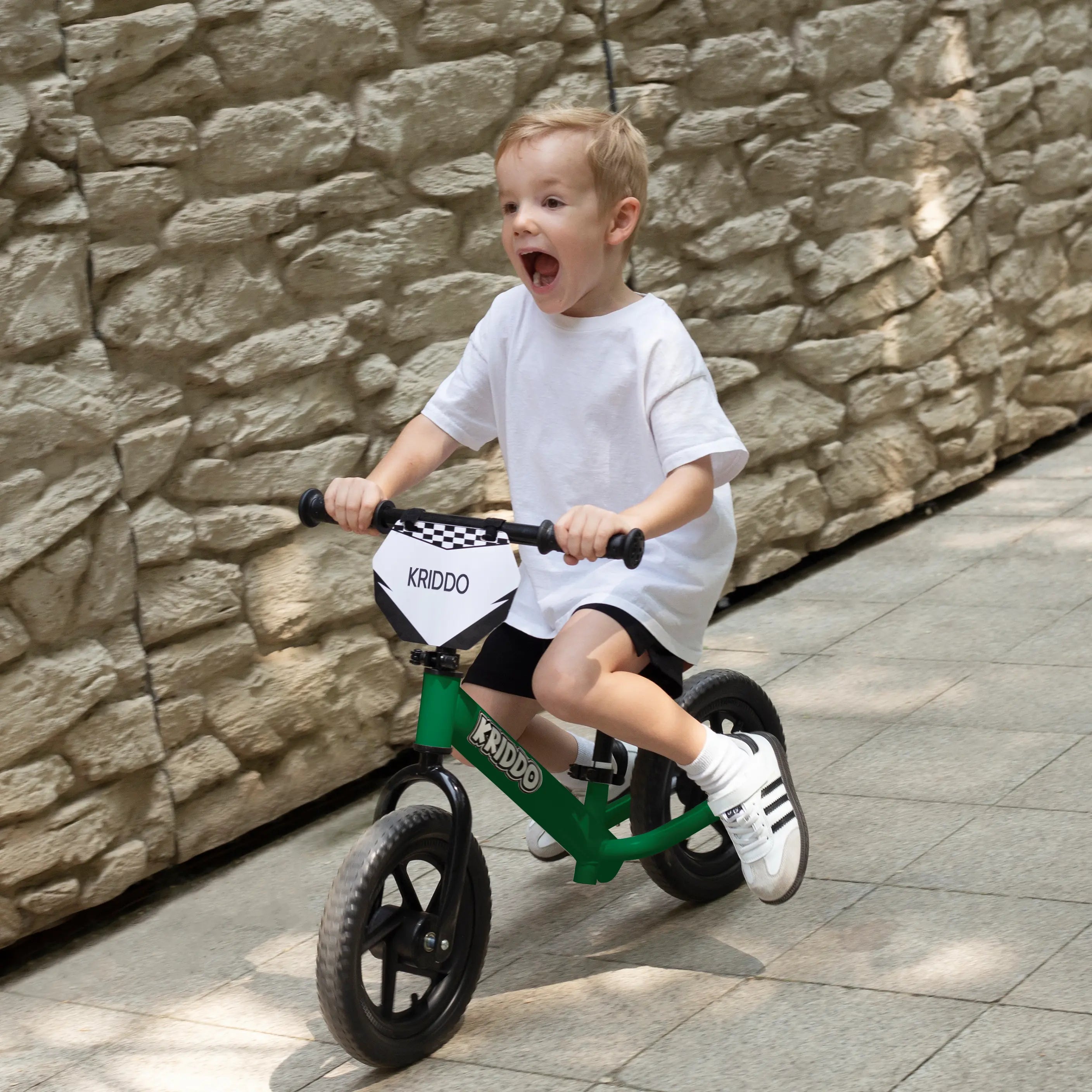 Child riding KRIDDO 12 Inch green balance bike against a stone wall.