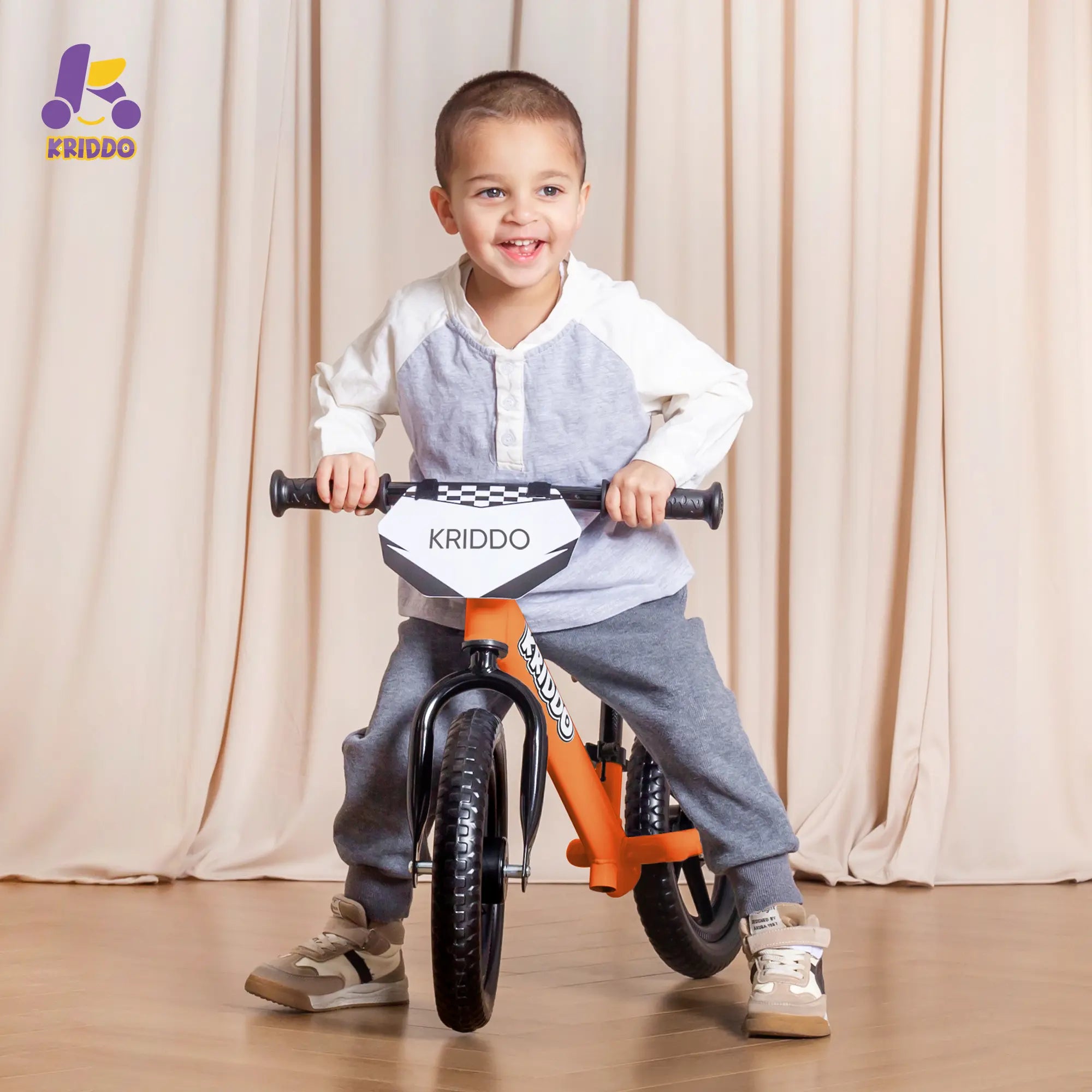 Toddler boy riding KRIDDO 12 Inch orange balance bike against a beige curtain