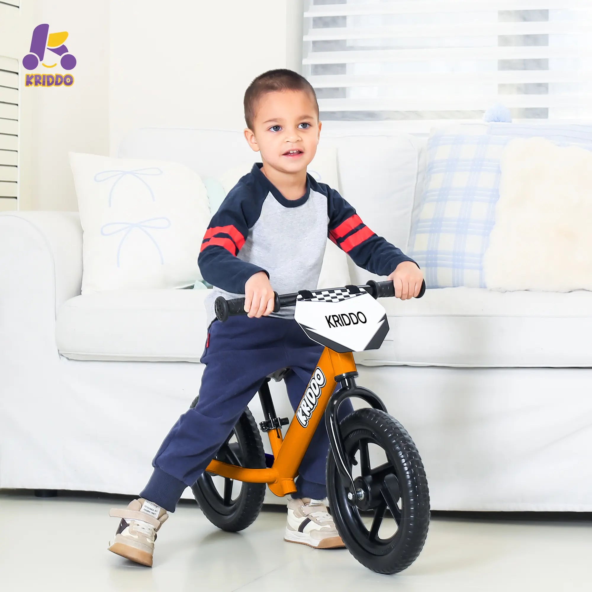 Child sitting on KRIDDO 12 Inch balance bike near by a sofa