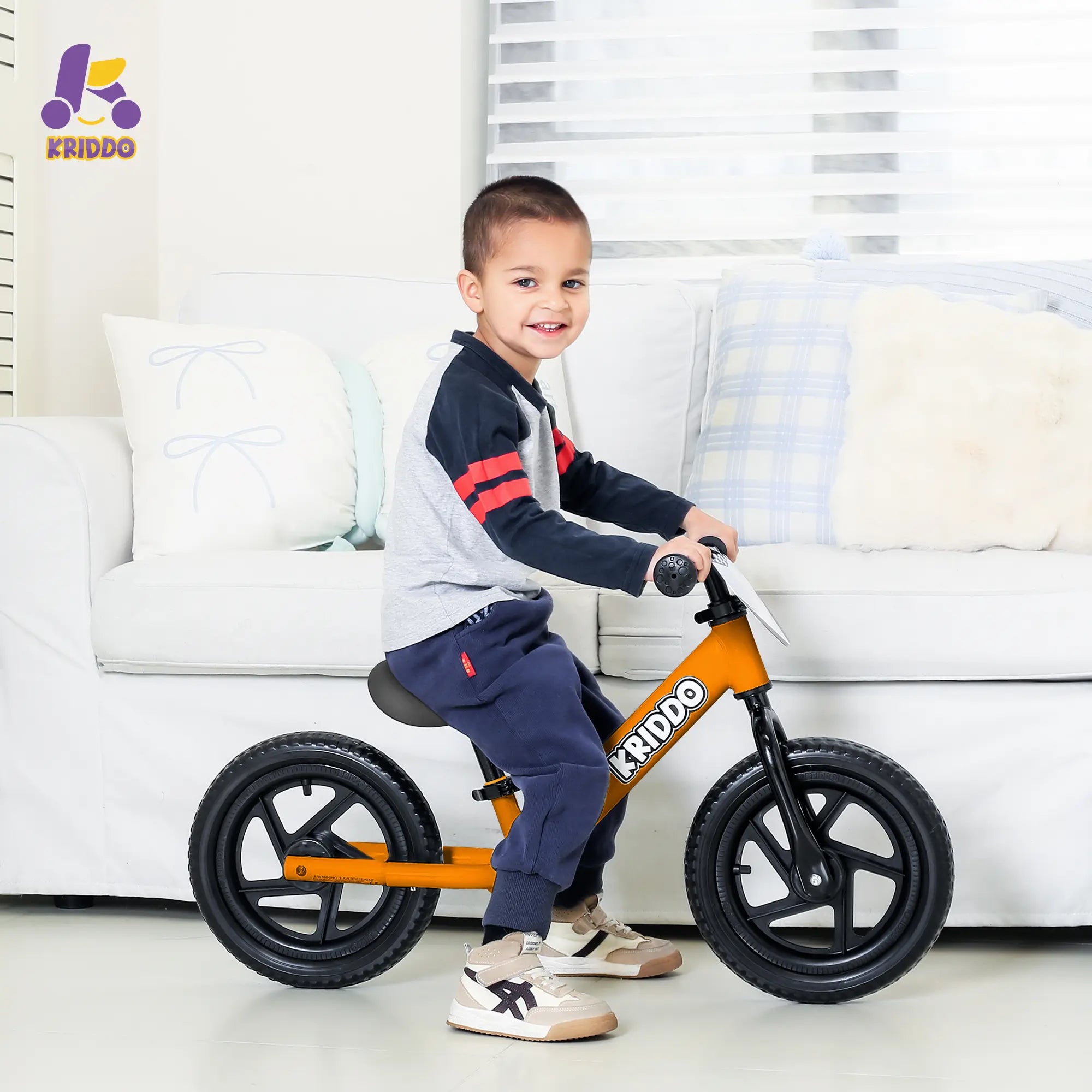 Boy riding KRIDDO Orange 12 Inch balance bike in a living room.