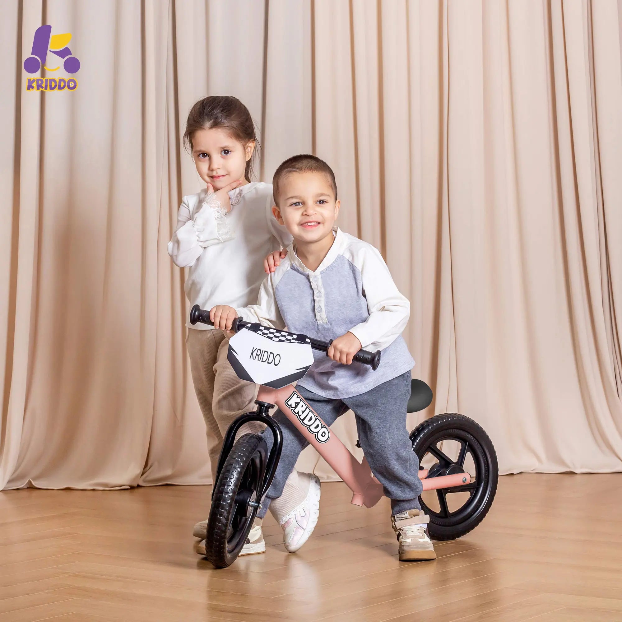 Two children with KRIDDO 12 inch balance bike in front of beige curtains