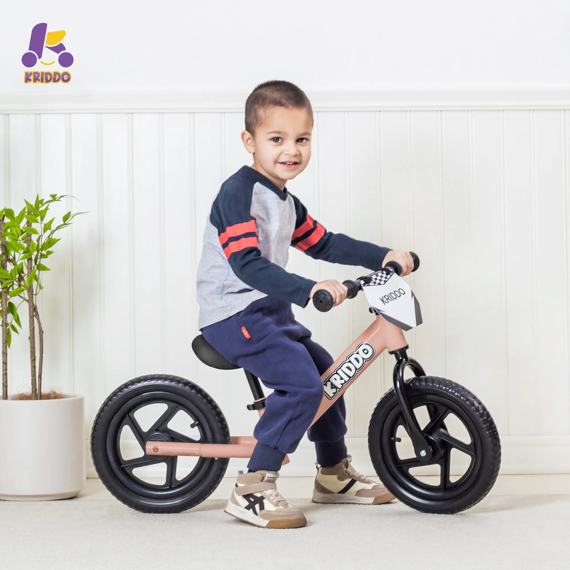 Boy riding KRIDDO Pink 12 Inch balance bike in a living room.