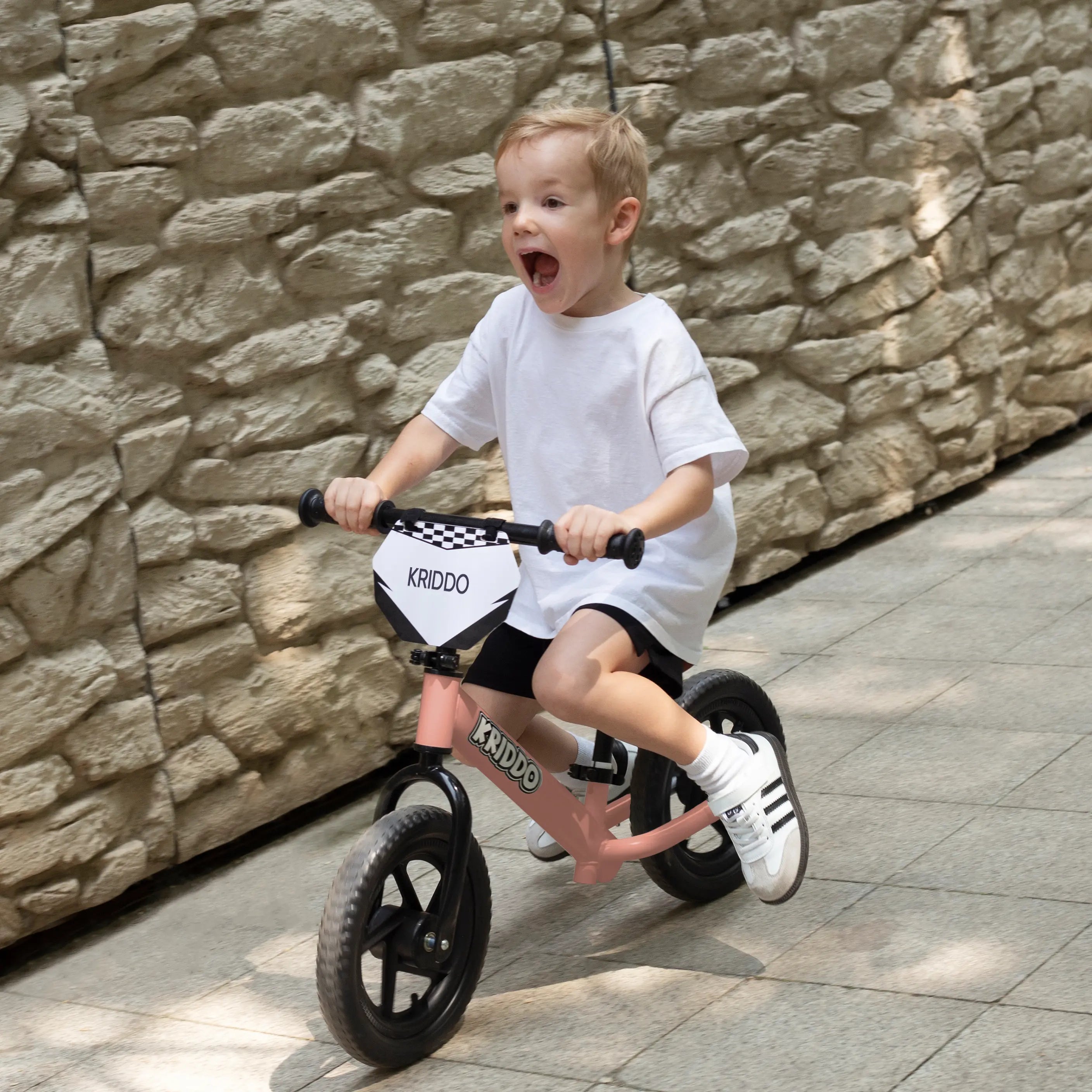 Little boy riding KRIDDO pink 12 inch balance bike against a stone wall