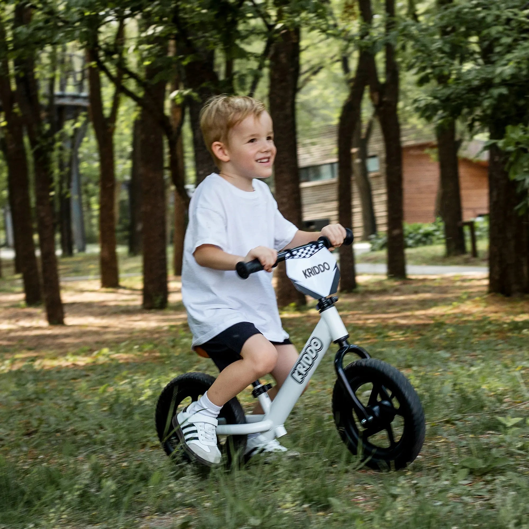 Child riding KRIDDO balance bike in a park with trees and grass.