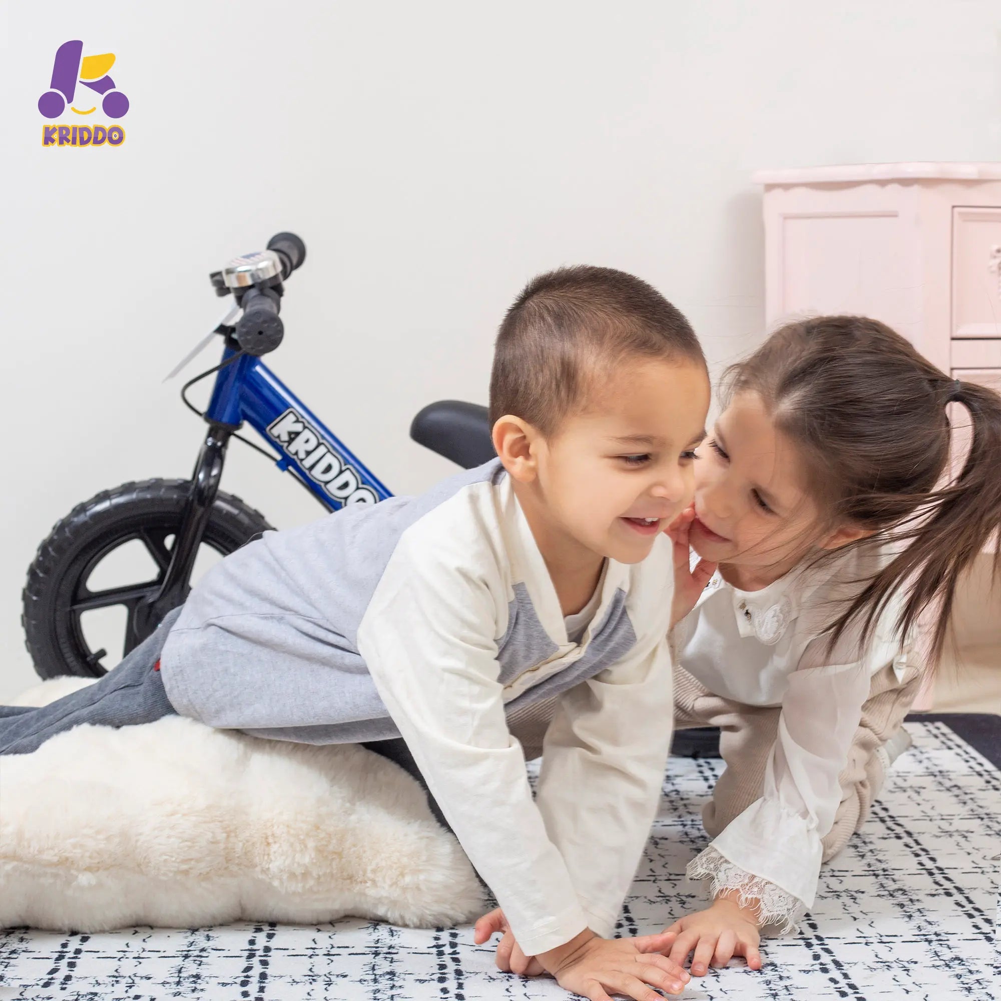 Two children playing on a bed with KRIDDO balance bike in the background