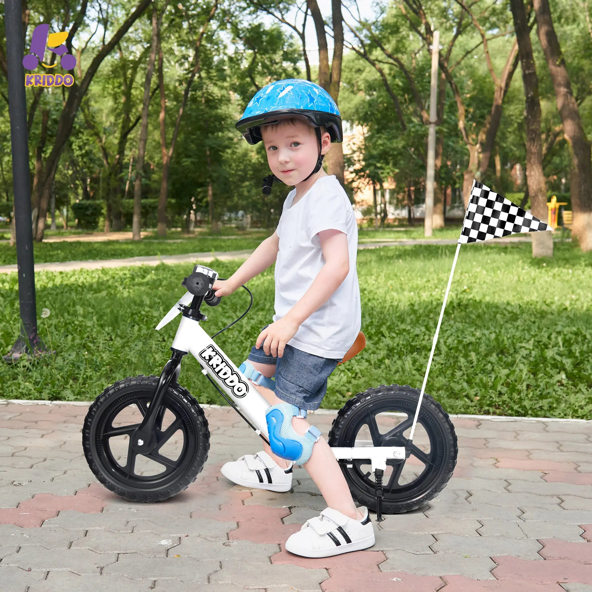Toddler boy riding a white balance bike with hand brake and a flag in a park, wearing a helmet.