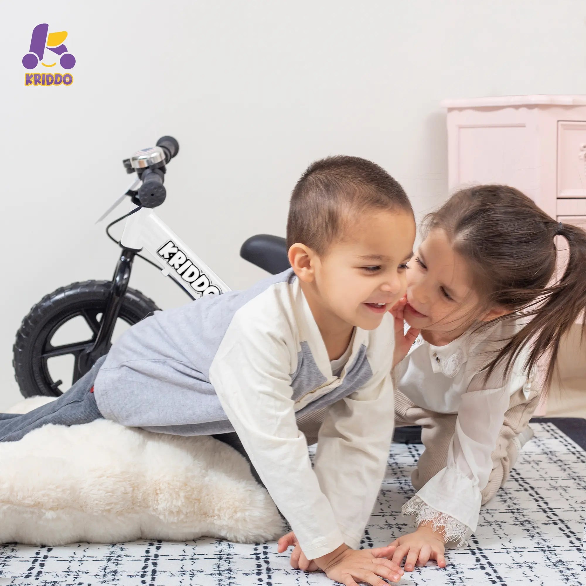 Two children playing on a bed with KRIDDO balance bike in the background