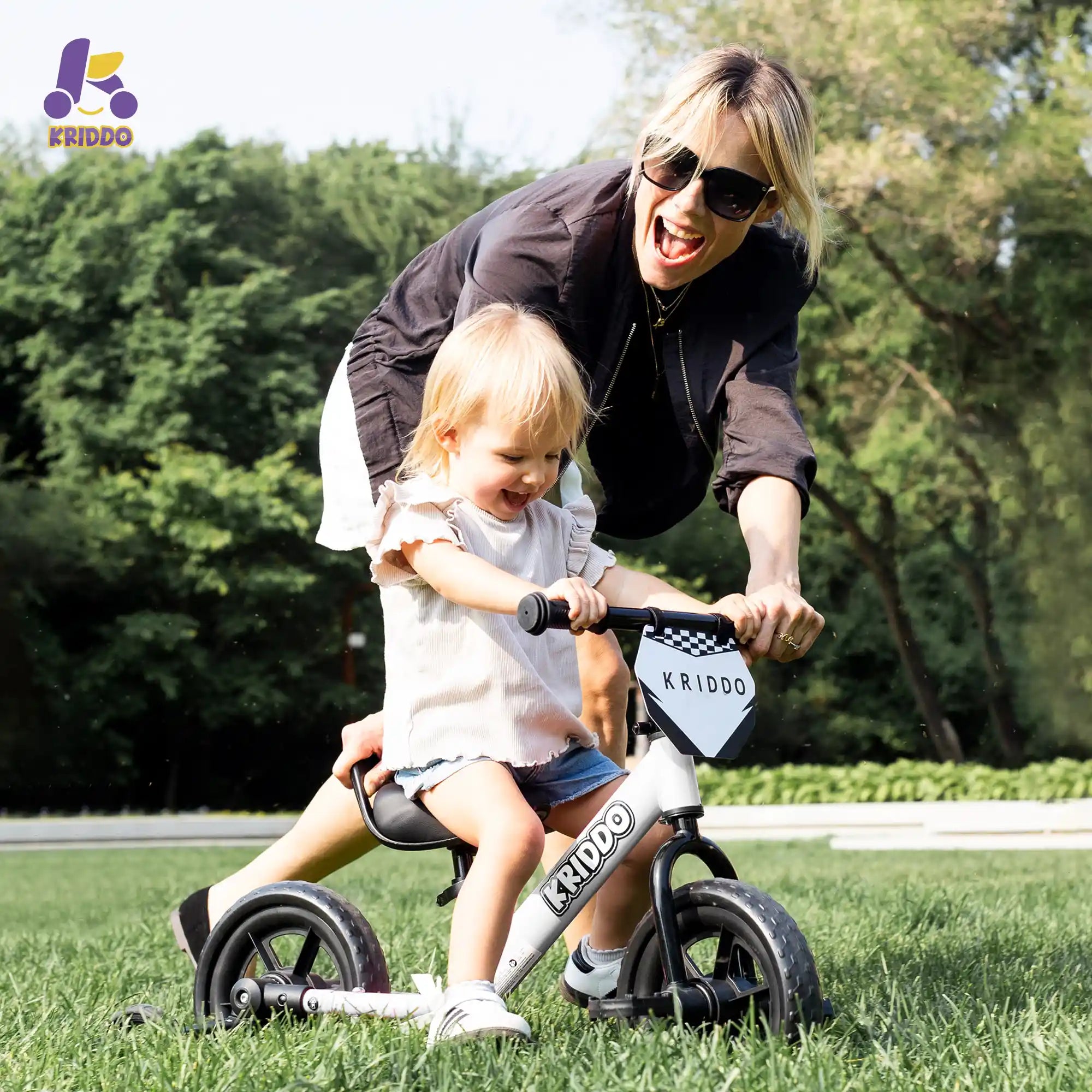 Woman assisting daughter on KRIDDO white convertible balance bike in a park.