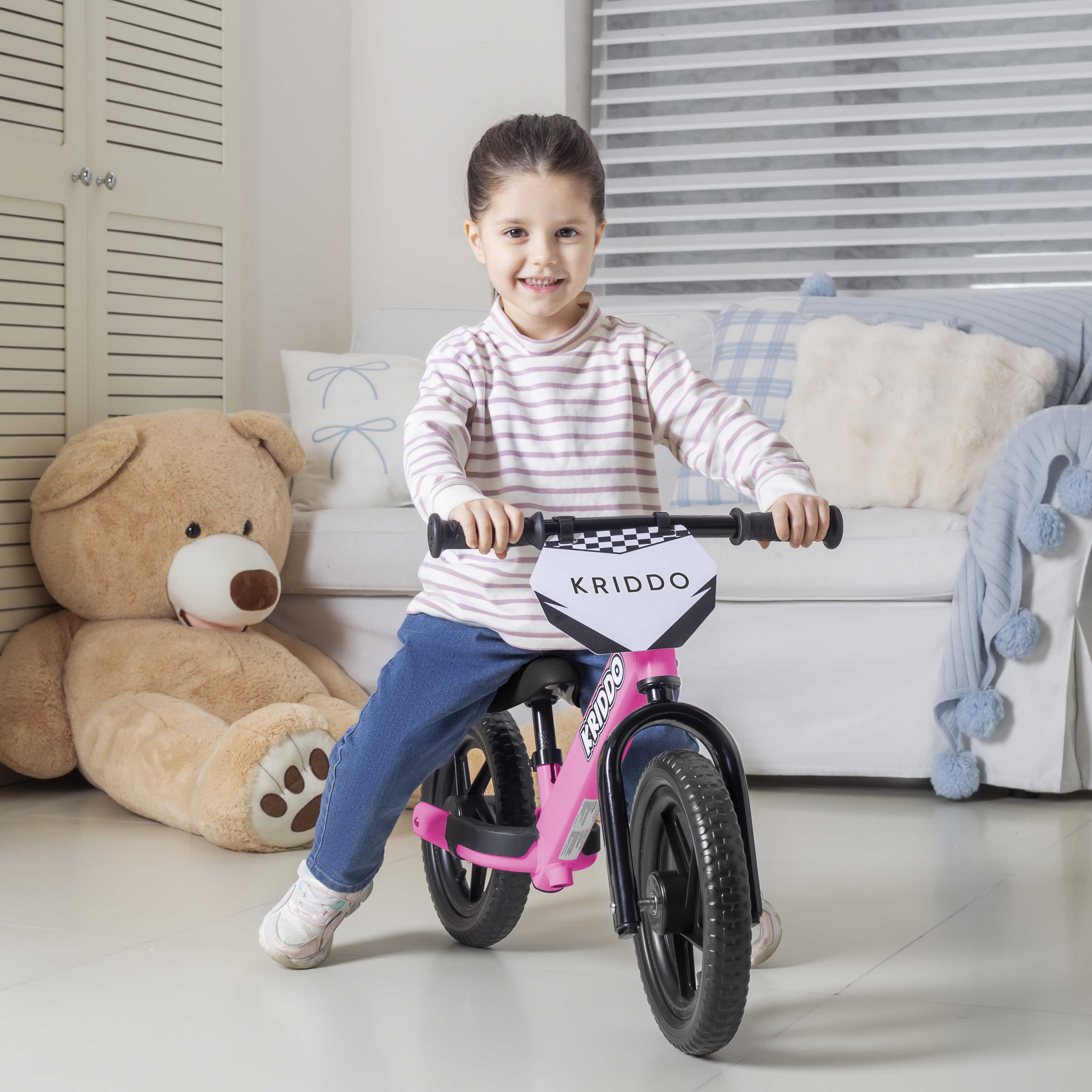 Girl riding KRIDDO pink balance bike in a living room with a teddy bear and sofa.