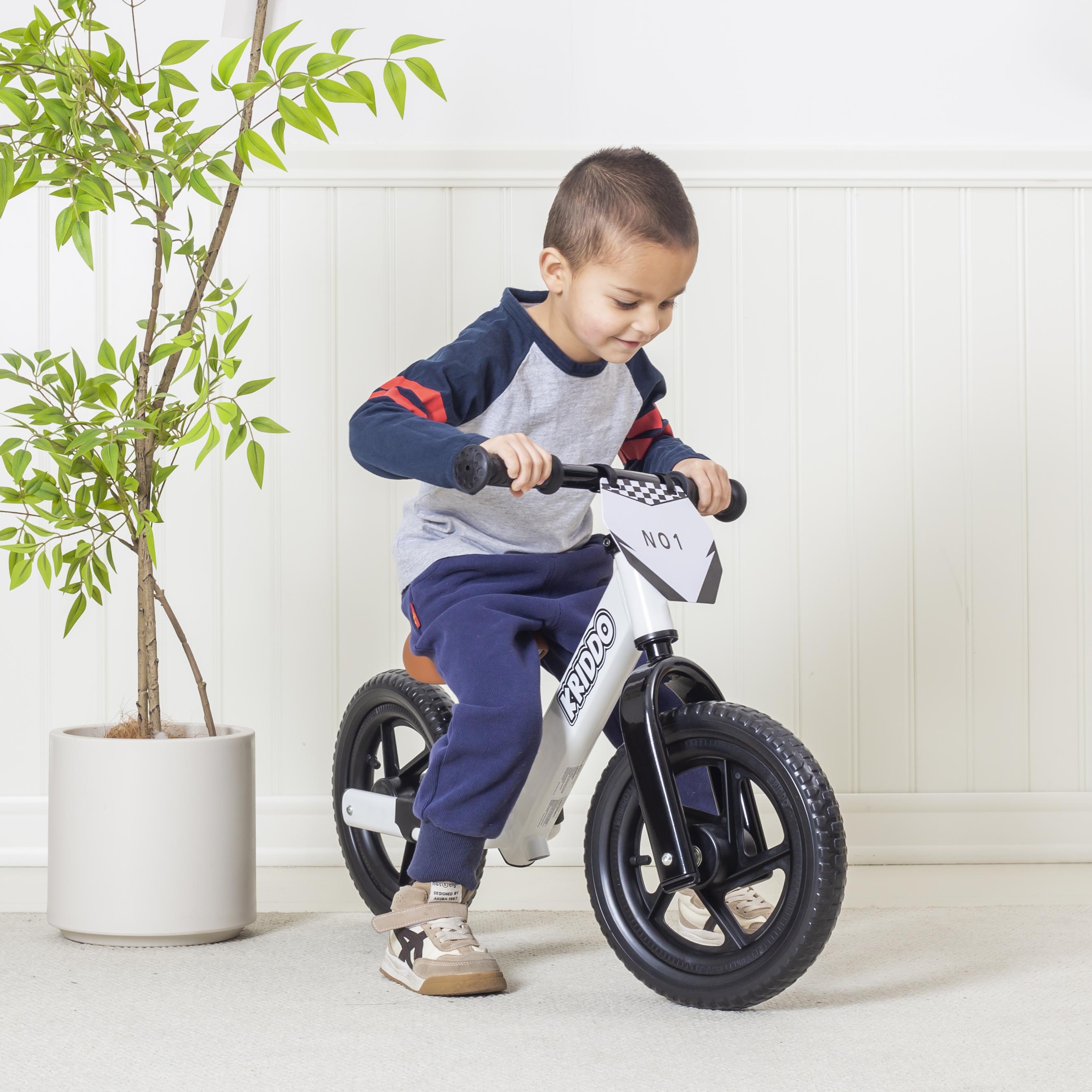 Boy riding a white balance bike indoors with a plant