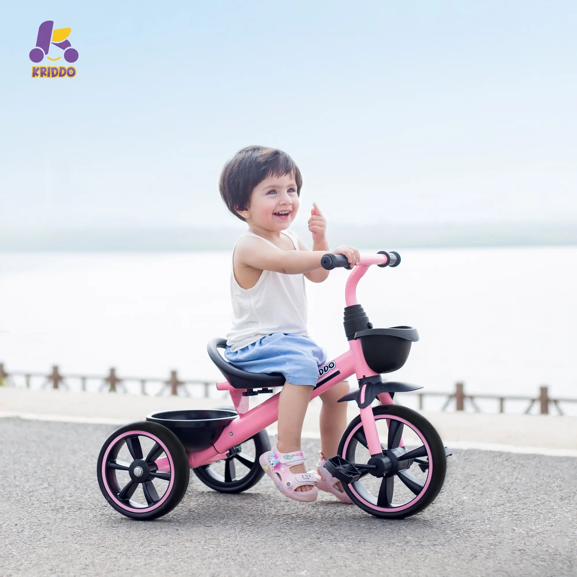 A toddler riding a pink KRIDDO tricycle by the lake.