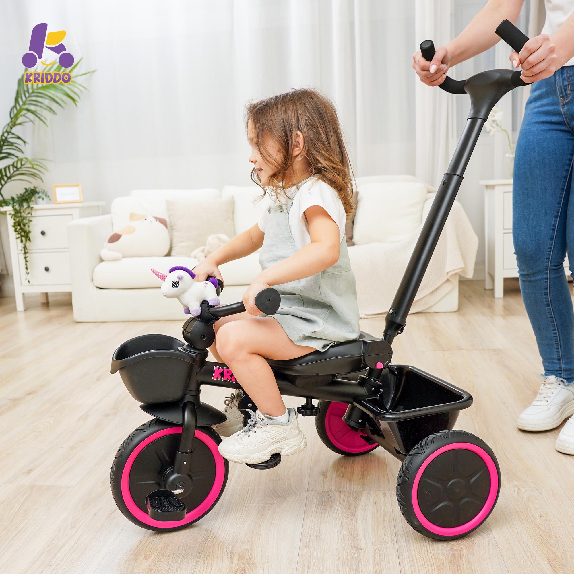 Child riding KRIDDO tricycle stroller with pink wheels in a living room, accompanied by an adult.