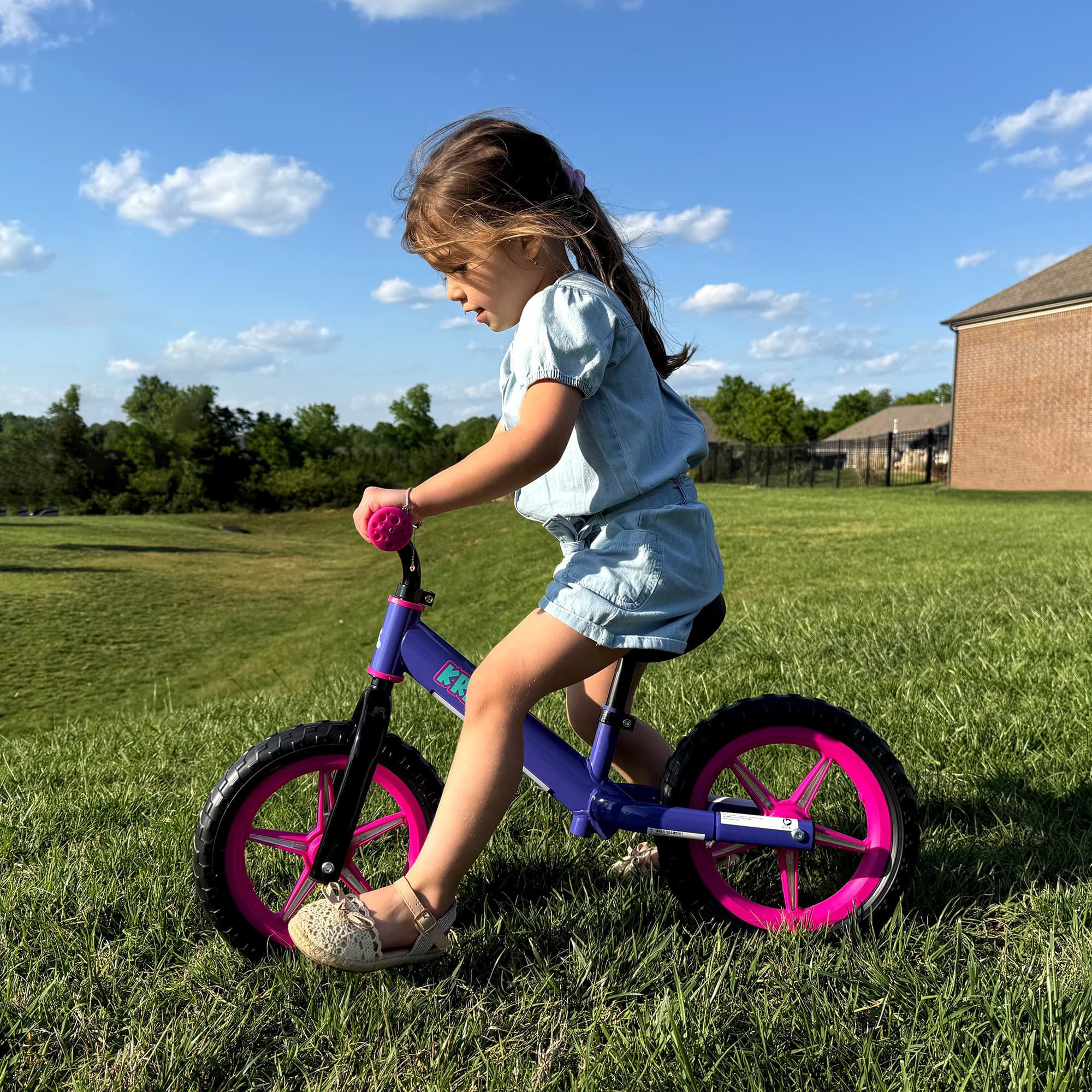 Girl riding KRIDDO light up balance bike in a grassy area with a clear sky