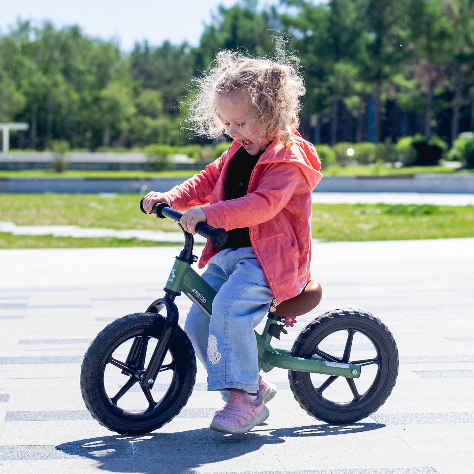 Child riding KRIDDO green lightweight balance bike on a paved path with trees