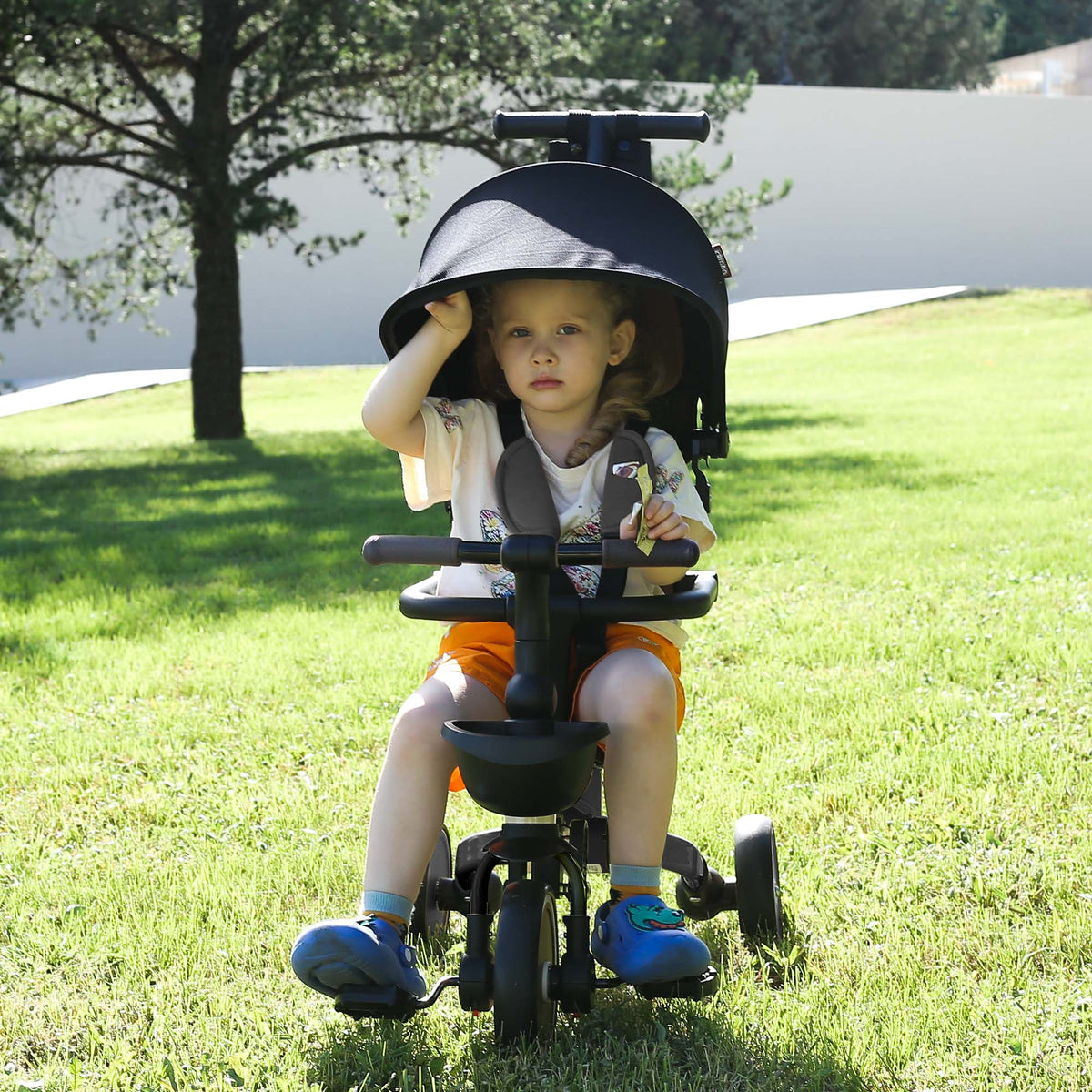 A toddler sits on KRIDDO foldable stroller trike outdoors on the grass
