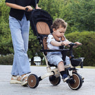 A toddler sits on KRIDDO 13 in 1 tricycle stroller, being pushed by an adult in a park. 