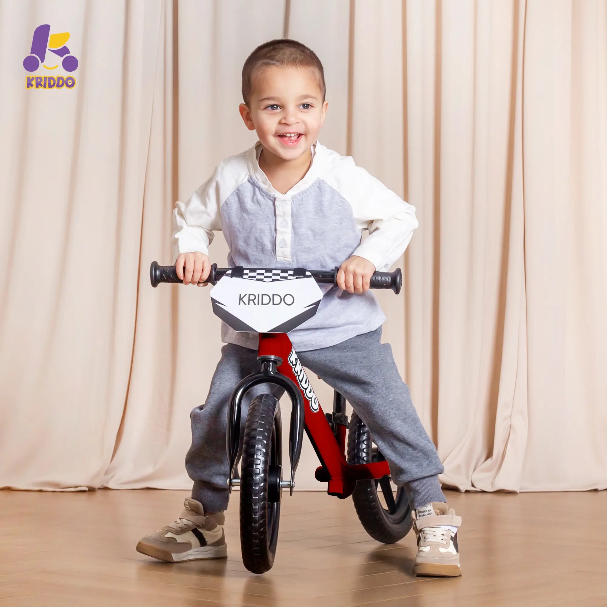 Toddler boy riding KRIDDO 12 Inch red balance bike against a beige curtain