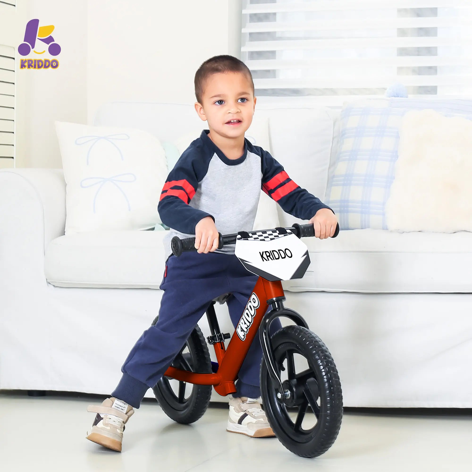 Child sitting on KRIDDO 12 Inch red balance bike near by a sofa