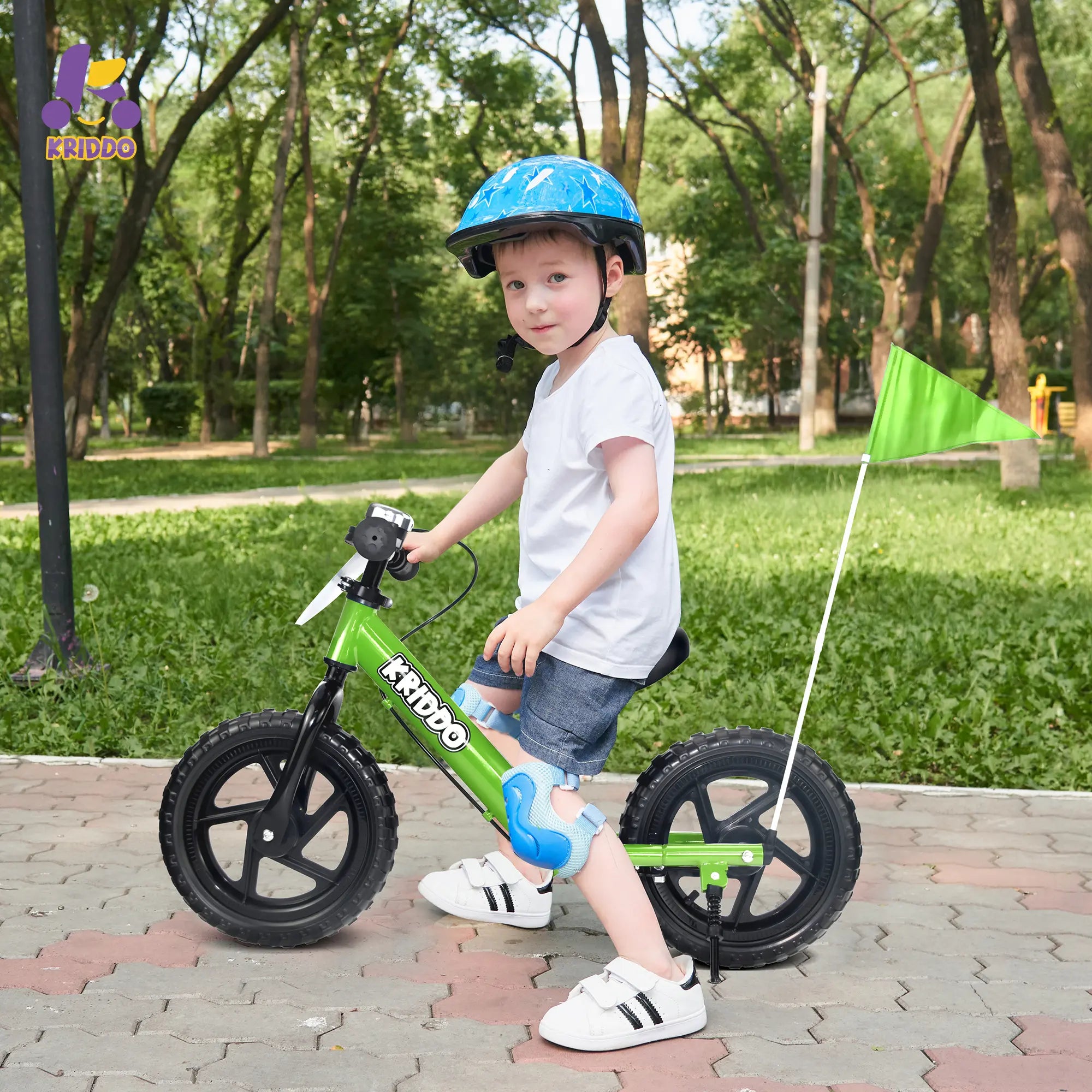 Toddler boy riding a green balance bike with hand brake and a flag in a park, wearing a helmet.