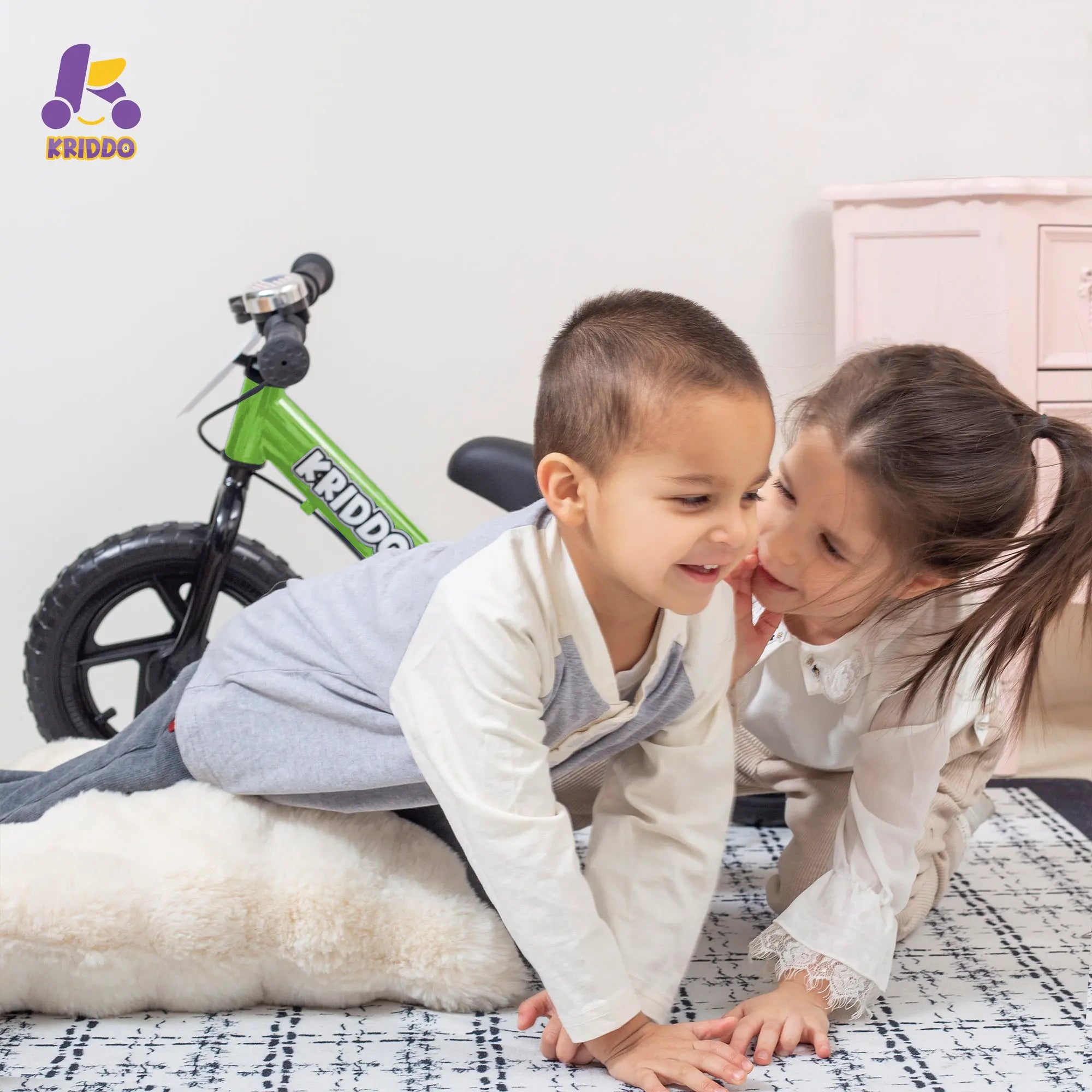 Two children playing on a bed with KRIDDO balance bike in the background