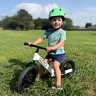 Toddler boy riding KRIDDO 12 inch balance bike in a grassy field with a helmet on