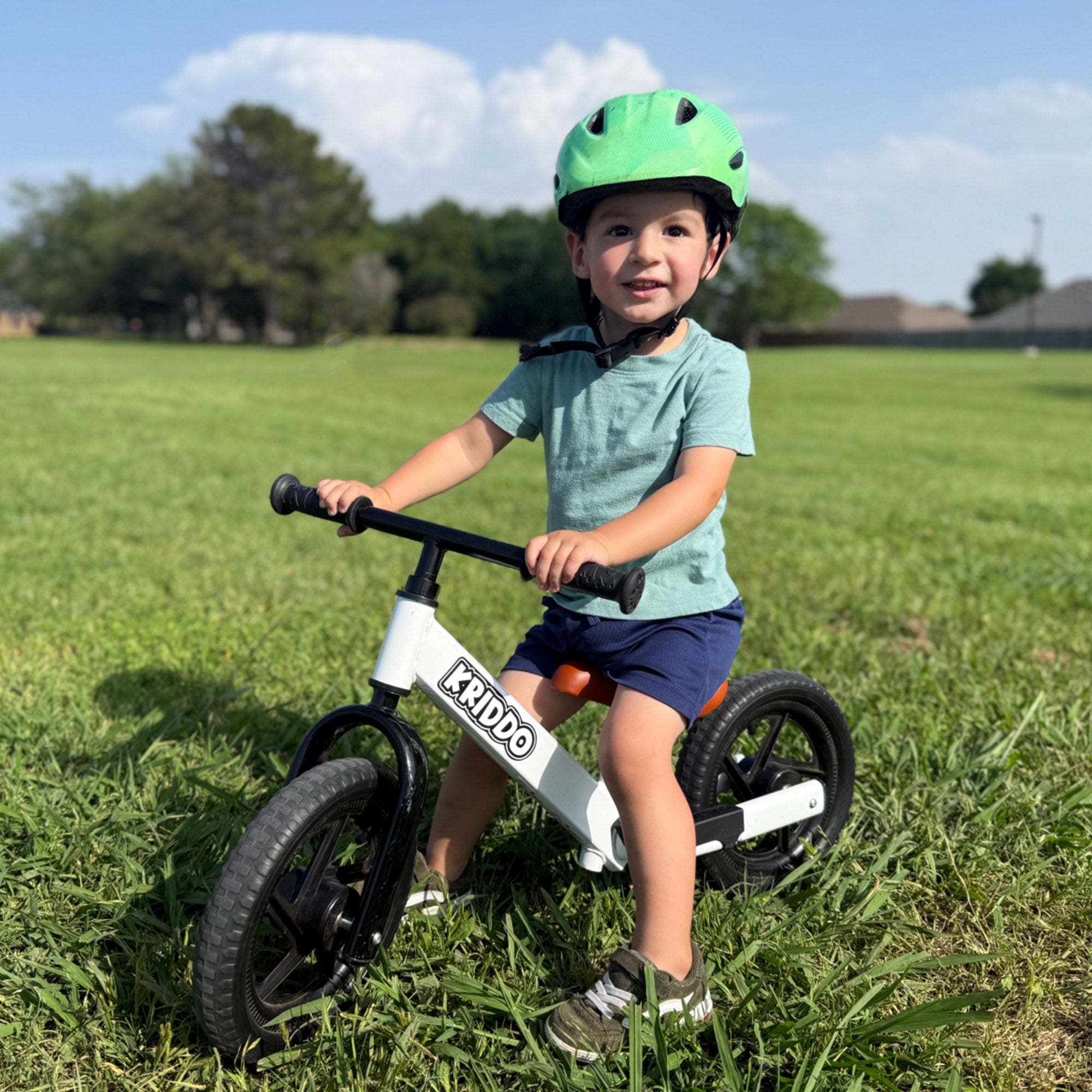 Toddler boy riding KRIDDO 12 inch balance bike in a grassy field with a helmet on