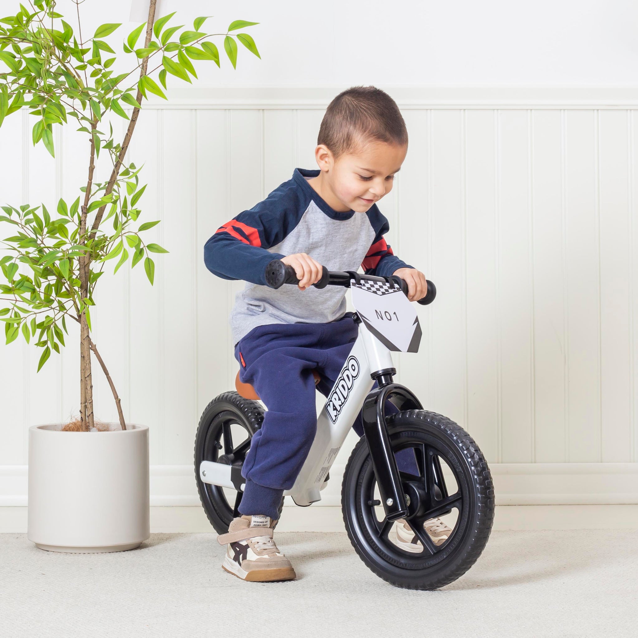 Boy riding a white balance bike indoors with a plant