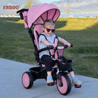 Little girl wearing heart-shaped sunglasses sits on KRIDDO light up tricycle stroller with a canopy in a park.