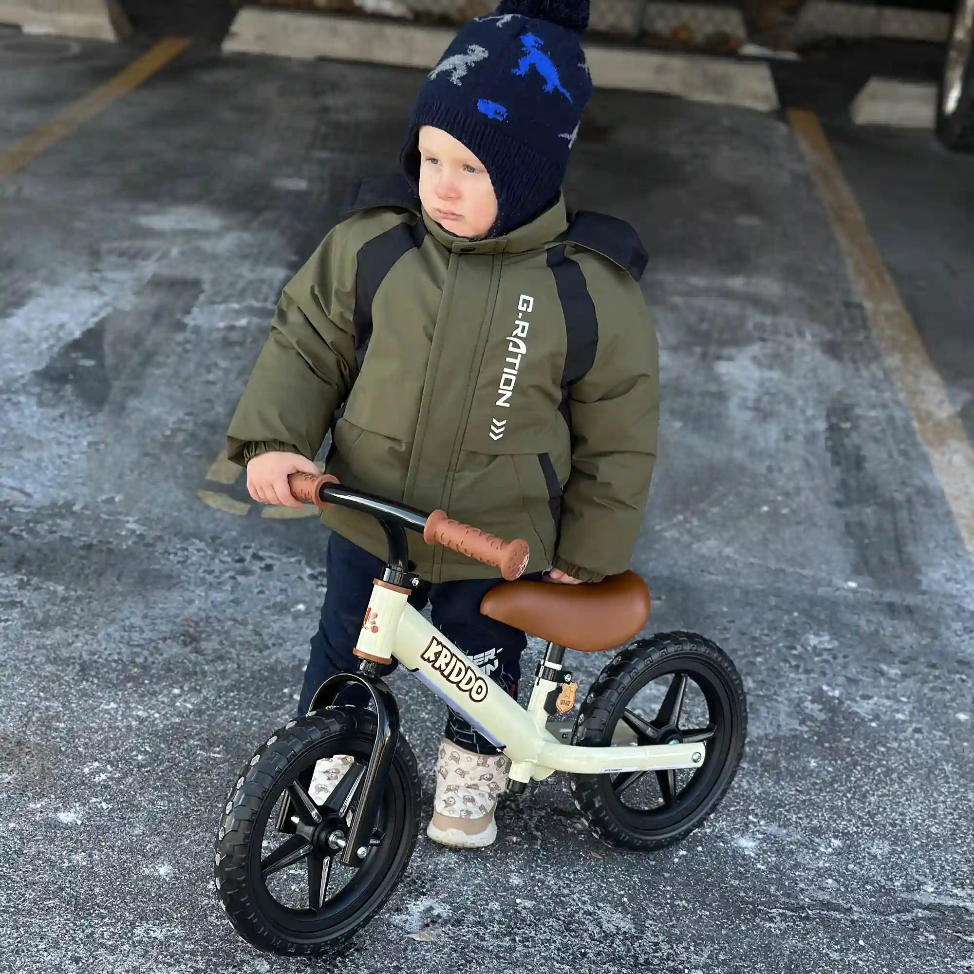 Child in a green jacket and blue beanie standing with KRIDDO light up balance bike on a concrete surface.