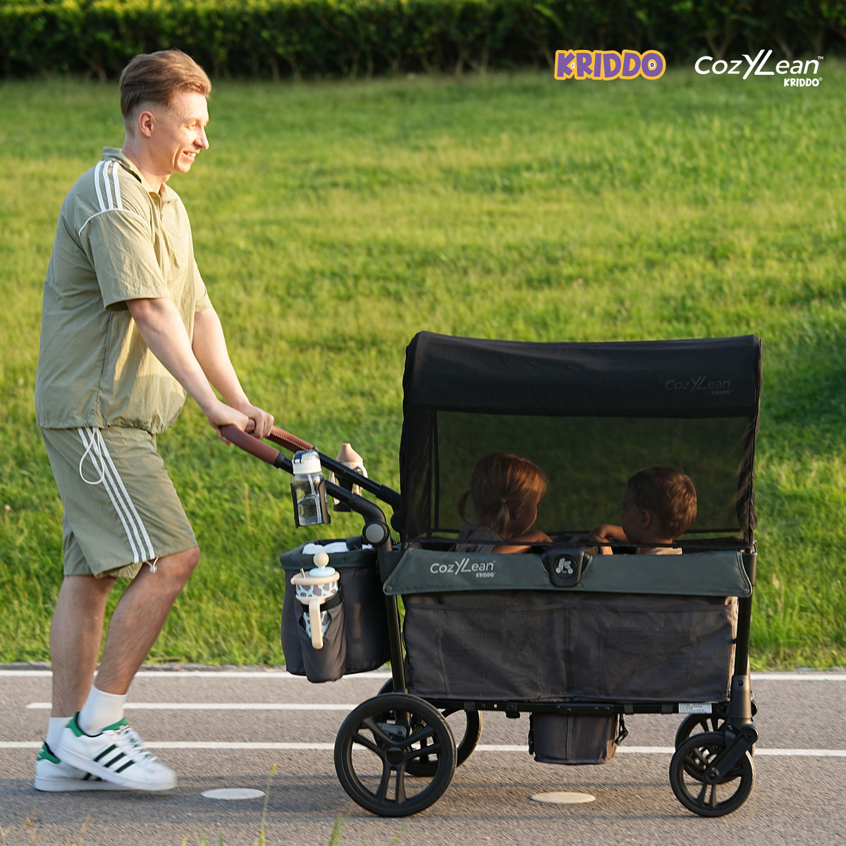 A man pushes KRIDDO CozyLean stroller with two children inside on a paved path in a grassy park.